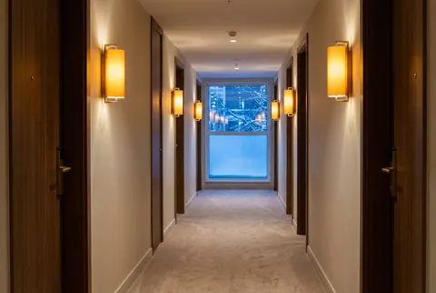 A quiet hotel corridor in the evening, warm wall lamps between wooden doors and a snowy blue window at the end