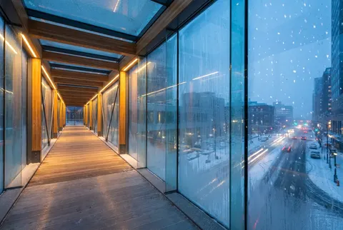 Inside a warm-lit glass pedestrian bridge over a snowy downtown street at dusk, frost on the glass