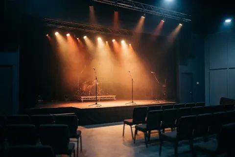 An empty show room stage with a drum riser and microphone stands under warm amber lights, rows of empty chairs in front